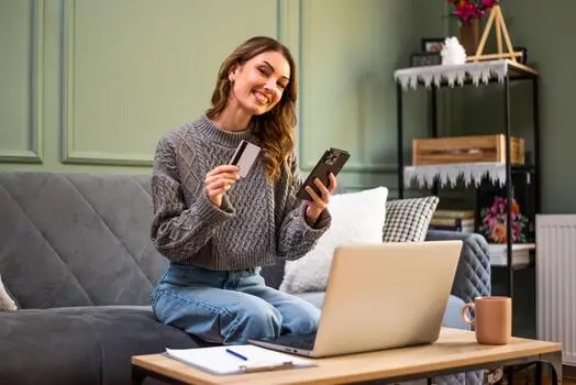 A woman smiling on the sofa at home, a card and her phone in her hands and a laptop open on the table.